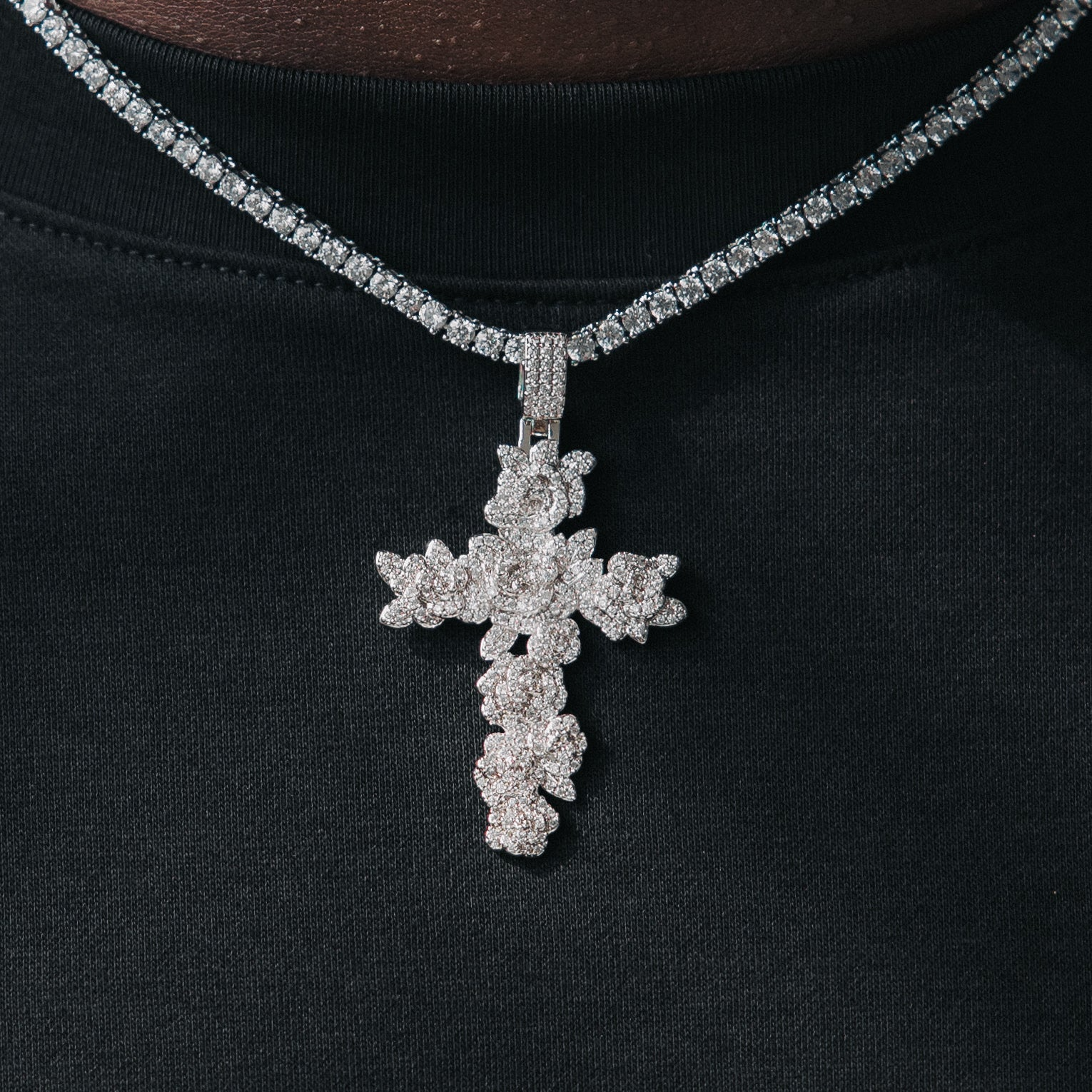 A person wearing a black hoodie with a white gold cross necklace featuring iced-out roseblades, hand-plucked diamonds, and a solid gold frame.