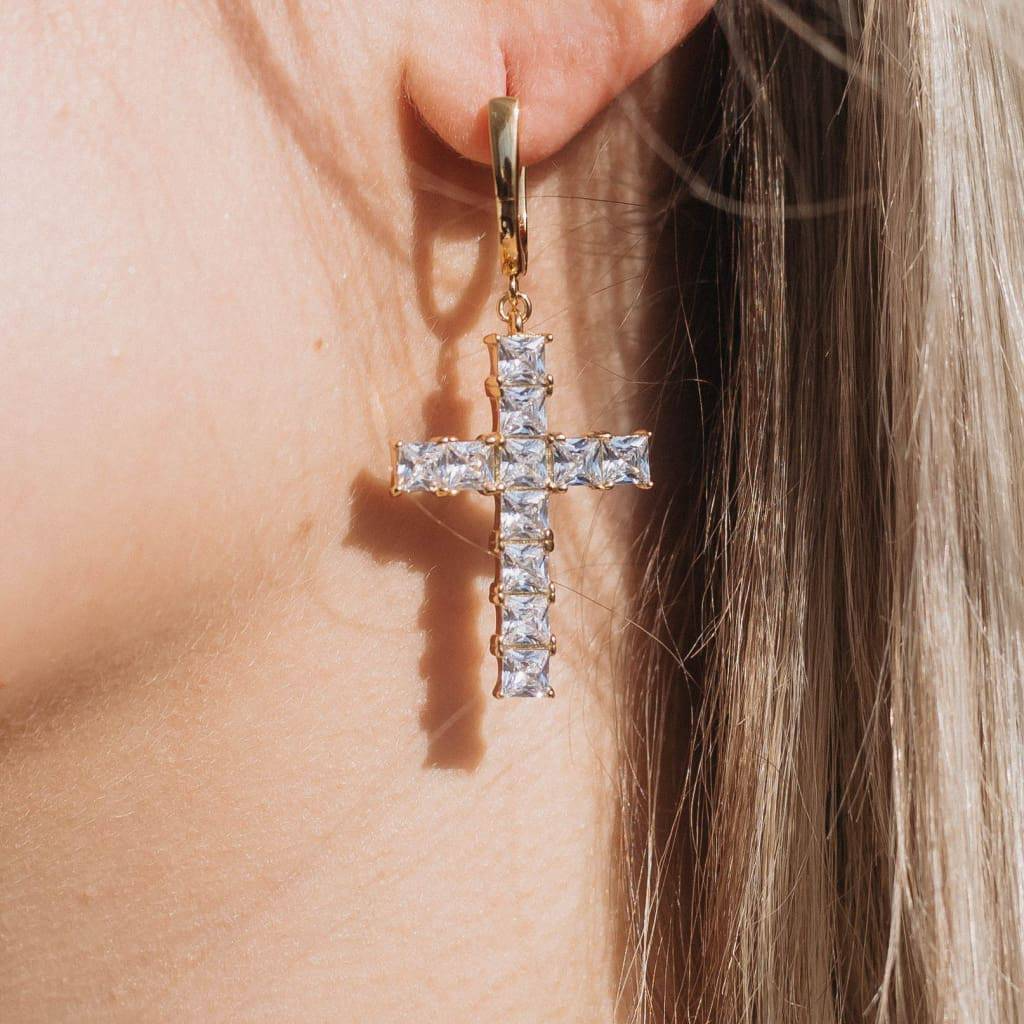 Close-up image of a person wearing gold cross-shaped earrings embedded with clear stones.