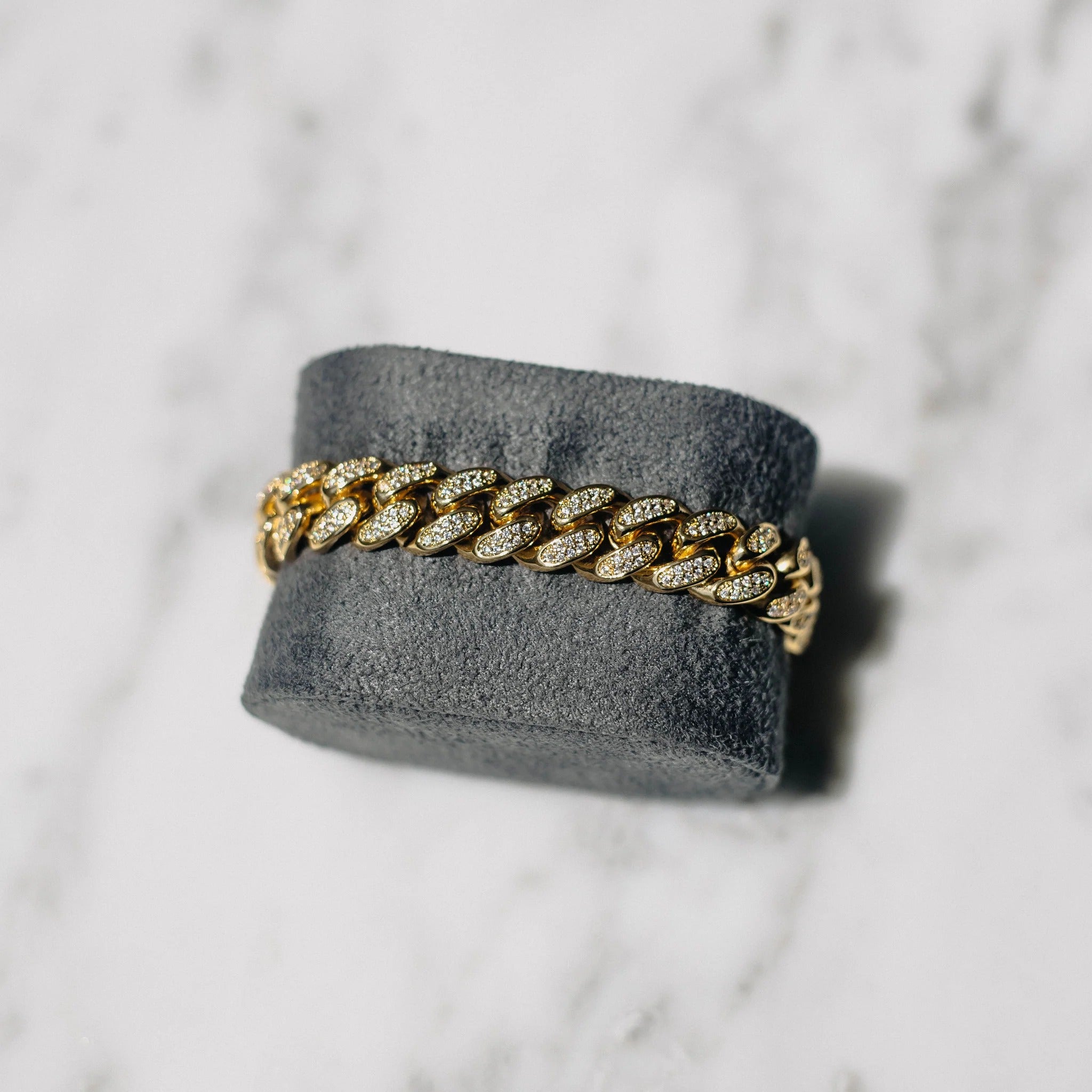 A gold-colored Cuban link chain bracelet displayed on a gray stand against a marble background.