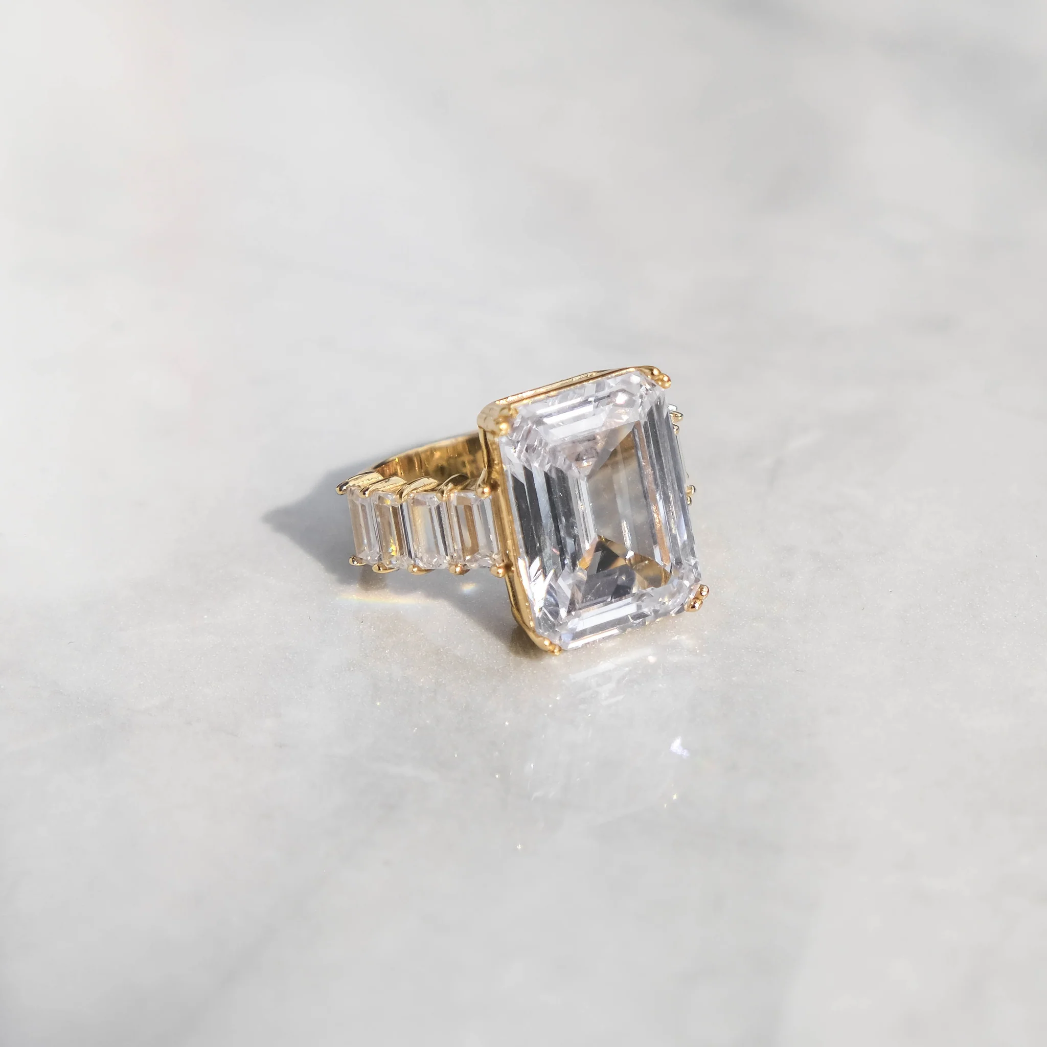 An 18K gold ring with a large emerald cut clear cubic zirconia centerpiece and smaller clear cubic zirconia accents on the band, displayed on a marble surface.